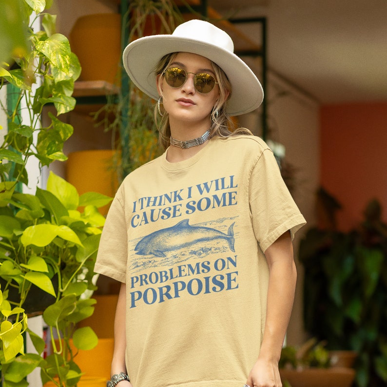May include: A light beige t-shirt with a blue graphic of a dolphin and the text "I think I will cause some problems on porpoise".