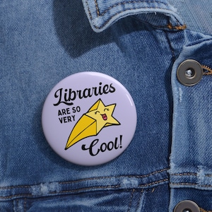 May include: A circular, pale purple badge with the words "Libraries ARE SO VERY Cool!" in black script. A yellow shooting star with a smiling face is in the centre. The badge is pinned to a blue denim jacket.