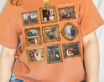 May include: A burnt orange t-shirt featuring framed reproductions of Edvard Munch paintings. The shirt displays nine different artworks, including "The Scream." The artist's name is printed in cursive on the upper right.
