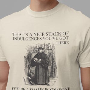 May include: A beige t-shirt with a black graphic print of a man in a robe holding a book. The text on the shirt reads "That's a nice stack of indulgences you've got there. It'd be a shame if someone wrote 95 theses about them. Martin Luther, probably 1517."