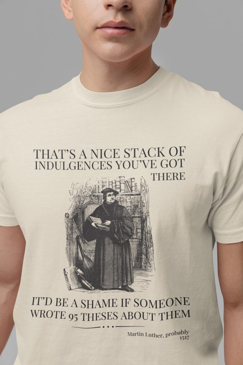 May include: A beige t-shirt with a black graphic print of a man in a robe holding a book. The text on the shirt reads "That's a nice stack of indulgences you've got there. It'd be a shame if someone wrote 95 theses about them. Martin Luther, probably 1517."