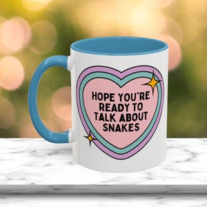 May include: White ceramic mug with a blue handle and interior. The mug features a heart-shaped design with the text "HOPE YOU'RE READY TO TALK ABOUT SNAKES" in black. The heart is outlined in pink, teal, and purple, with gold star accents.