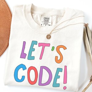 Op de afbeelding: Crèmekleurig T-shirt met de tekst "LET'S CODE!" in kleurrijke letters. Een gouden ketting met een hanger ligt op het shirt. Het shirt is opgevouwen en heeft een label bij de halslijn.
