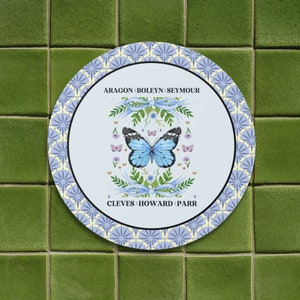 Puede incluir: Un plato redondo blanco con un patr&oacute;n azul y blanco. El centro del plato presenta una mariposa azul con un borde blanco, rodeada de flores y cintas. El texto "ARAGON - BOLEYN - SEYMOUR" est&aacute; por encima de la mariposa, y el texto "CLEVES - HOWARD - PARR" est&aacute; por debajo de la mariposa.
