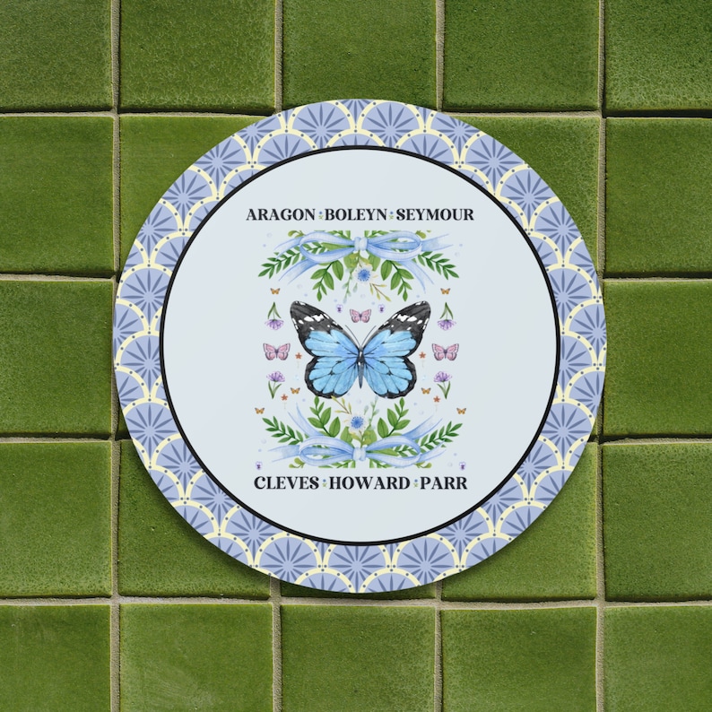 Puede incluir: Un plato redondo blanco con un patr&oacute;n azul y blanco. El centro del plato presenta una mariposa azul con un borde blanco, rodeada de flores y cintas. El texto "ARAGON - BOLEYN - SEYMOUR" est&aacute; por encima de la mariposa, y el texto "CLEVES - HOWARD - PARR" est&aacute; por debajo de la mariposa.