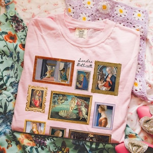 May include: Pink t-shirt with a graphic print of Sandro Botticelli's paintings, including "The Birth of Venus" and "The Annunciation".