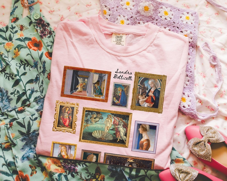 Puede incluir: Camiseta rosa con un estampado gr&aacute;fico de pinturas de Sandro Botticelli, incluyendo "El nacimiento de Venus" y "La Anunciaci&oacute;n".