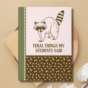 May include: A hardback notebook with a tan cover and a green spine. The front features a raccoon illustration and the text "FERAL THINGS MY STUDENTS SAID" in brown. The bottom border has a pattern of small white flowers on a brown background.
