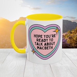 May include: White ceramic mug with a yellow interior and handle. The mug features a heart-shaped design with the text "HOPE YOU'RE READY TO TALK ABOUT MACBETH" in a pink and blue color scheme. Two gold starbursts accent the design.