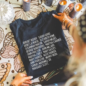 Puede incluir: Camiseta negra con texto blanco que enumera los nombres de los acusados en los juicios de brujas de Salem. El texto dice: Bridget Bishop, Sarah Good, Rebecca Nurse, Elizabeth Howe, Susannah Martin, Sarah Wildes, George Burroughs, Martha Carrier, John Proctor, John Willard, Martha Corey, Mary Easty, Mary Parker, Alice Parker, Ann Pudeator, Wilmot Redd, Samuel Wardwell Sr., Margaret Scott.