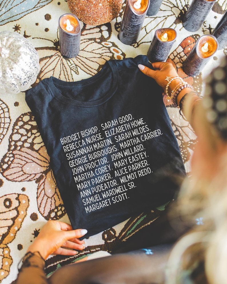 Puede incluir: Camiseta negra con texto blanco que enumera los nombres de los acusados en los juicios de brujas de Salem. El texto dice: Bridget Bishop, Sarah Good, Rebecca Nurse, Elizabeth Howe, Susannah Martin, Sarah Wildes, George Burroughs, Martha Carrier, John Proctor, John Willard, Martha Corey, Mary Easty, Mary Parker, Alice Parker, Ann Pudeator, Wilmot Redd, Samuel Wardwell Sr., Margaret Scott.