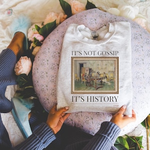 May include: A grey sweatshirt with the text "It's not gossip, it's history" and a framed image of a painting depicting a group of people gathered around a table. The painting is in a vintage style.