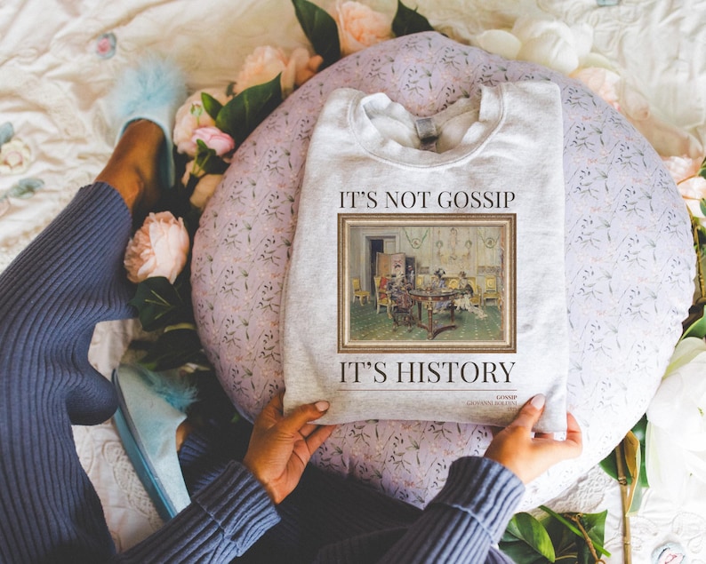 May include: A grey sweatshirt with the text "It's not gossip, it's history" and a framed image of a painting depicting a group of people gathered around a table. The painting is in a vintage style.