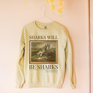 May include: A light yellow sweatshirt with a painting of a shark attack on a boat. The text "SHARKS WILL BE SHARKS" is printed on the sweatshirt. The painting is titled "Watson and the Shark" by John Singleton Copley, 1778.