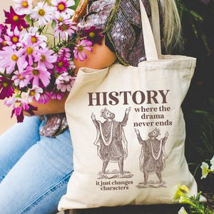 May include: Beige tote bag with the word "HISTORY" and the phrase "where the drama never ends" printed in brown. The bag features illustrations of two figures in historical garb. The phrase "it just changes characters" is printed below.