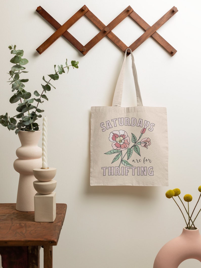 May include: A beige canvas tote bag with the text "Saturdays are for thrifting" in pink and green lettering. A pink flower with green leaves is printed on the bag.