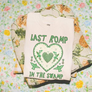 May include: Cream-colored t-shirt with the text "LAST ROMP IN THE SWAMP" in green, featuring a green heart design with floral and bird accents. The shirt is laid on a floral patterned pillow.