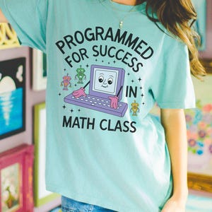 May include: A light blue t-shirt with the text "PROGRAMMED FOR SUCCESS IN MATH CLASS" in black. The graphic features a smiling computer with pink hands and two robot characters. The shirt is worn with blue denim shorts.