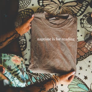 K&ouml;nnte beinhalten: Ein gefaltetes braunes T-Shirt mit dem wei&szlig;en Text "naptime is for reading". Das Shirt wird von H&auml;nden mit Armb&auml;ndern gehalten und liegt auf einer Oberfl&auml;che mit Schmetterlings- und Blumenmustern. Zwei brennende Kerzen sind zu sehen.