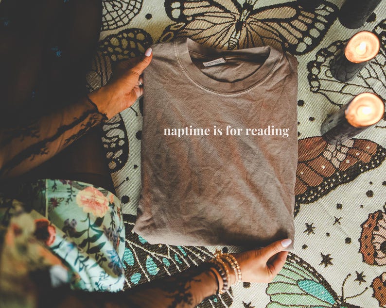 K&ouml;nnte beinhalten: Ein gefaltetes braunes T-Shirt mit dem wei&szlig;en Text "naptime is for reading". Das Shirt wird von H&auml;nden mit Armb&auml;ndern gehalten und liegt auf einer Oberfl&auml;che mit Schmetterlings- und Blumenmustern. Zwei brennende Kerzen sind zu sehen.
