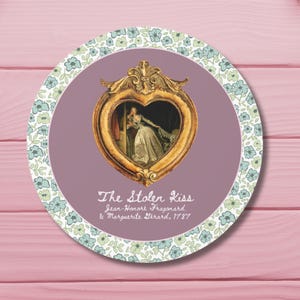 Puede incluir: Posavasos redondo con un marco dorado en forma de corazón que contiene una pintura de una mujer con un vestido blanco. El fondo es de color púrpura apagado. El posavasos tiene el texto "The Stolen Kiss" y motivos florales.