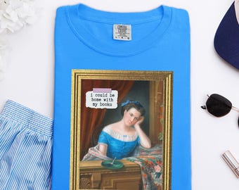 Könnte beinhalten: Ein leuchtend blaues T-Shirt mit einem gerahmten Bild einer Frau in einem blauen Kleid, die ihren Kopf auf ihre Hand stützt. Das Bild enthält den Text "I could be home with my books". Das T-Shirt ist ordentlich gefaltet.