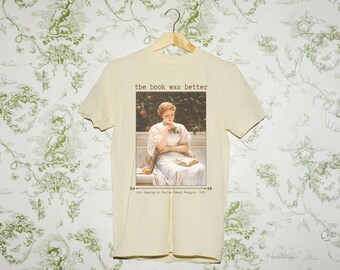 Puede incluir: Camiseta color crema con una imagen enmarcada de una mujer leyendo, con el texto "the book was better" encima. Debajo de la imagen está el texto "Girl Reading by Charles Howard, 1900". La camiseta está colgada en una percha de madera.