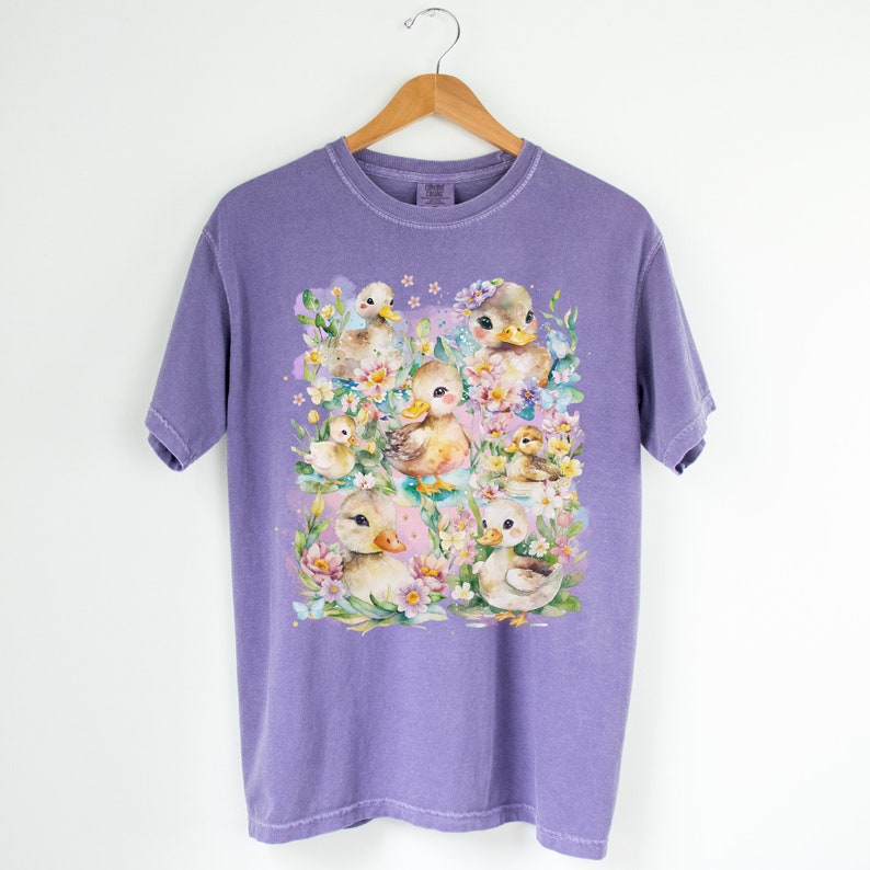 Puede incluir: Camiseta morada con un dise&ntilde;o de acuarela de un grupo de patitos rodeados de flores.