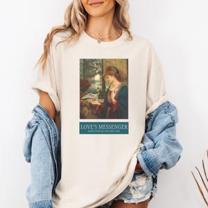 May include: Cream-colored t-shirt featuring a framed print of a painting titled "Love's Messenger" by Marie Spartali Stillman. The artwork depicts a woman in a blue and red dress with a dove. The text "LOVE'S MESSENGER" is printed in white on a teal banner.