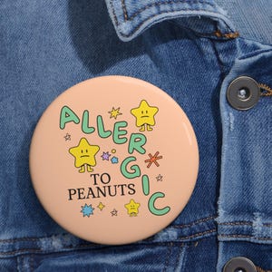 Può includere: Un bottone color pesca con il testo "ALLERGIC TO PEANUTS" in lettere verdi e nere. Il testo è circondato da stelle gialle con facce e altre stelle colorate.
