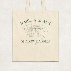 May include: Cream-colored tote bag with green text and illustrations. The text reads "RAISE A GLASS TO SHADOW DADDIES" above an illustration of three cocktail glasses being clinked. The bag has two handles.