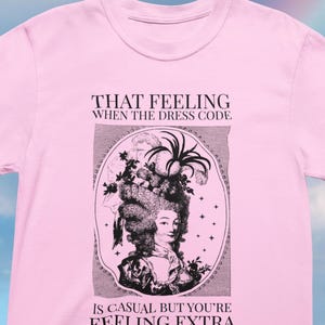 Könnte beinhalten: Ein hellrosa T-Shirt mit kurzen Ärmeln und einem schwarzen Grafikdruck einer Figur aus dem 18. Jahrhundert mit aufwendiger Perücke und Hut. Der Text lautet: THAT FEELING WHEN THE DRESS CODE IS CASUAL BUT YOU'RE FEELING EXTRA, zentriert über und unter dem Bild.