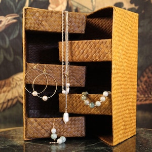 May include: A woven jewellery organiser with multiple compartments, displaying various pieces of jewellery. Gold hoop earrings with pearls, a silver necklace with a pearl pendant, and a beaded bracelet are shown. The organiser is made of a light brown material.