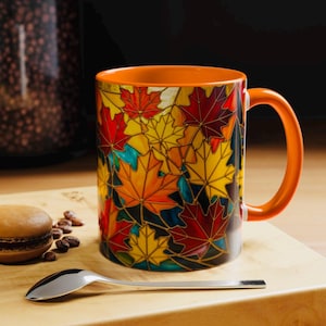 May include: A ceramic mug with an orange handle and interior. The exterior features a colorful stained-glass design of autumn maple leaves in shades of red, yellow, and orange. The mug sits on a wooden surface with a spoon and a pastry.