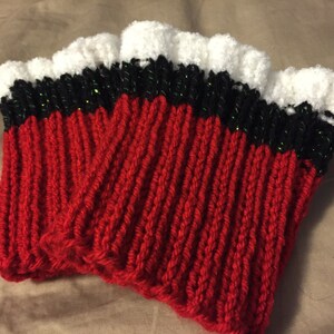Op de afbeelding: Een paar gebreide beenwarmers. De beenwarmers zijn voornamelijk rood met een zwarte en groene band en een witte, gerimpelde rand. Het ribgebreide ontwerp is zichtbaar. De beenwarmers worden op een neutrale achtergrond gepresenteerd.