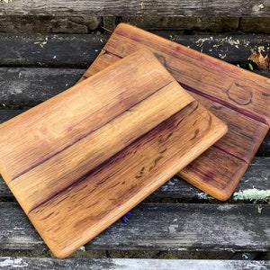 May include: Two rectangular wooden cutting boards with rounded edges. The boards have a natural wood grain pattern with reddish-brown and purple hues. The boards are displayed on a weathered wooden surface.