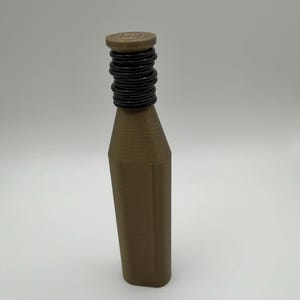 Puede incluir: Una botella marr&oacute;n con un cuello negro y acanalado y una parte superior plana y circular. La botella tiene forma rectangular con lados angulados. La parte superior de la botella tiene un peque&ntilde;o dise&ntilde;o cuadrado.