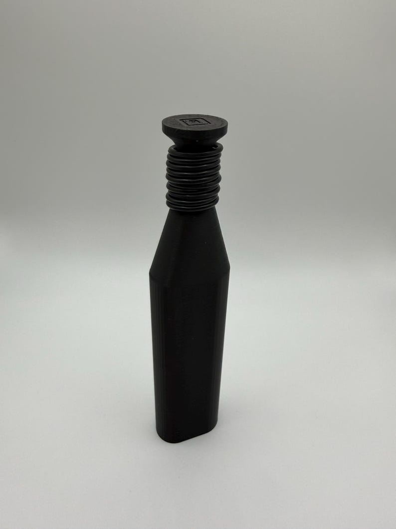 May include: A black bottle stopper with a cylindrical neck and a flat, round top. The neck features a series of horizontal ridges. The top has a square design in the center. The bottle stopper is made of a solid, matte black material.