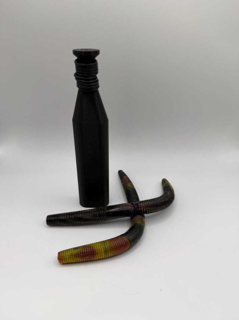 May include: A black bottle with a ridged neck and a tapered body stands next to two multi-colored fishing lures. The lures are curved and have a segmented design with shades of green, yellow, and red.