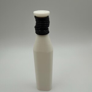 Puede incluir: Una botella blanca con un cuello negro enrollado y una tapa blanca. La botella tiene una base rectangular y un cuerpo ligeramente c&oacute;nico. La tapa parece tener un dise&ntilde;o circular. El fondo es de color neutro.