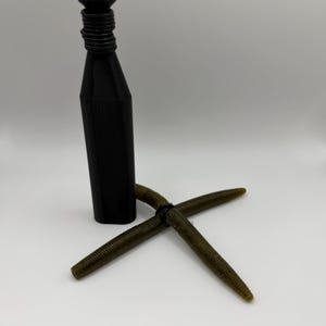 Puede incluir: Un tap&oacute;n de botella negro, cil&iacute;ndrico, con cuello acanalado, junto con dos se&ntilde;uelos de pesca verdes en forma de gusano. Los se&ntilde;uelos est&aacute;n cruzados y el tap&oacute;n est&aacute; en posici&oacute;n vertical. Los art&iacute;culos est&aacute;n sobre un fondo blanco.