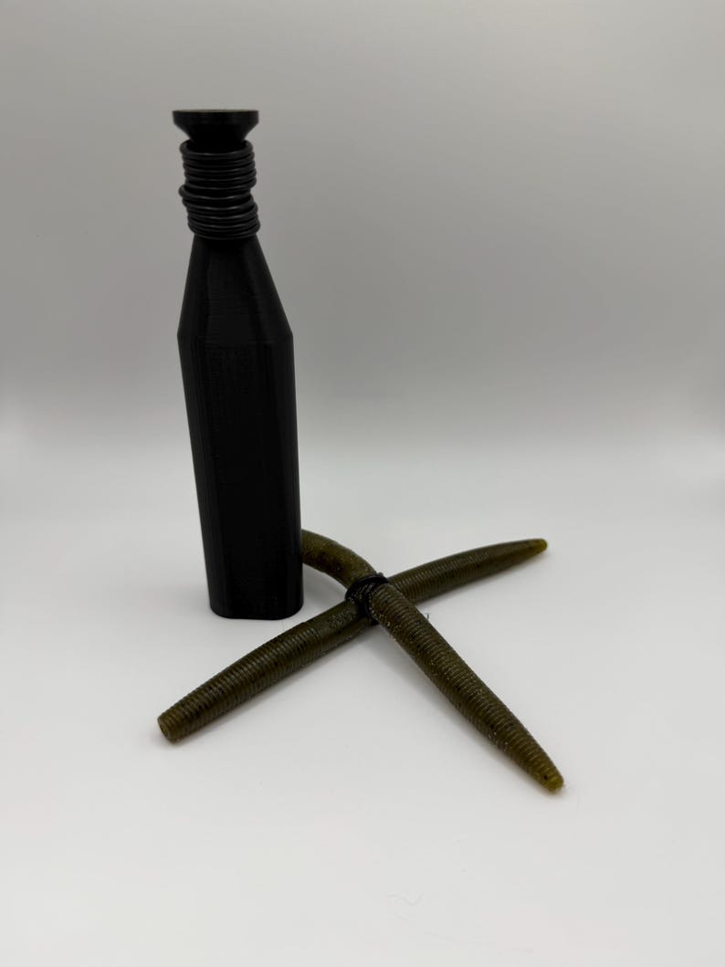 Puede incluir: Un tap&oacute;n de botella negro, cil&iacute;ndrico, con cuello acanalado, junto con dos se&ntilde;uelos de pesca verdes en forma de gusano. Los se&ntilde;uelos est&aacute;n cruzados y el tap&oacute;n est&aacute; en posici&oacute;n vertical. Los art&iacute;culos est&aacute;n sobre un fondo blanco.