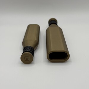 May include: Two brown bottle stoppers with black rubber seals. One stopper is inserted into a bottle, and the other is separate. The stoppers have a geometric design and a small square logo on the top. The background is plain white.