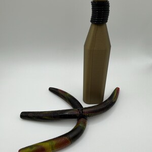 May include: A brown bottle with a black neck and a star-shaped fishing lure. The lure is black with red, yellow, and green accents. The bottle has a cylindrical neck and a flat top.