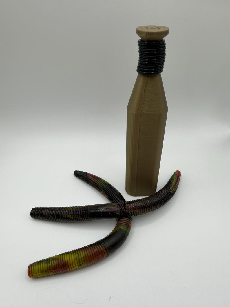 May include: A brown bottle with a black neck and a star-shaped fishing lure. The lure is black with red, yellow, and green accents. The bottle has a cylindrical neck and a flat top.