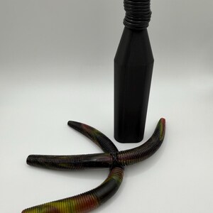 May include: A black bottle stopper with a ridged neck and a four-armed, multi-colored fishing lure. The lure is primarily black with red, yellow, and green accents. The bottle stopper is approximately 6 inches tall.