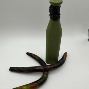 May include: An olive green bottle with a black neck and a matching cap. Three multi-colored fishing lures are arranged in front of the bottle. The lures are black, red, and yellow.