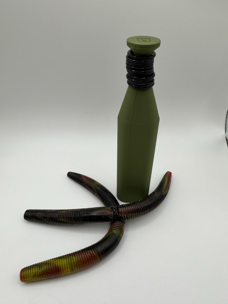 May include: An olive green bottle with a black neck and a matching cap. Three multi-colored fishing lures are arranged in front of the bottle. The lures are black, red, and yellow.