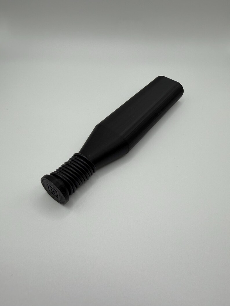 Puede incluir: Un tap&oacute;n de botella negro con una parte superior rectangular y una base acanalada. El tap&oacute;n tiene un cuerpo largo y rectangular y est&aacute; dise&ntilde;ado para encajar en una botella. El tap&oacute;n est&aacute; hecho de un material negro mate.
