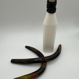 May include: A white bottle with a black ringed neck and a white top. Three dark brown, ribbed, curved fishing lures with red and green accents are arranged in front of the bottle.